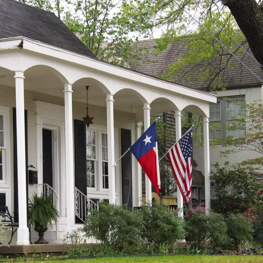 A white house with black shutters featuring a front porch displaying the Texas state flag and the American flag, with greenery and a tree in the foreground. Inside, homeowners can enjoy enhanced comfort with a top-of-the-line home water softener system.