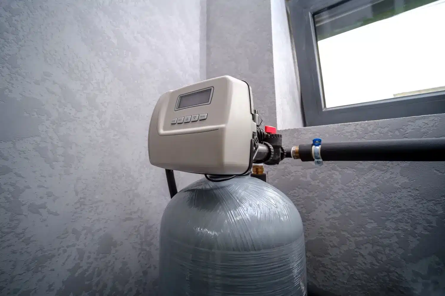 A home water softener unit installed indoors with pipes connected, positioned near a window, offers the myriad benefits of water softeners for your entire household.