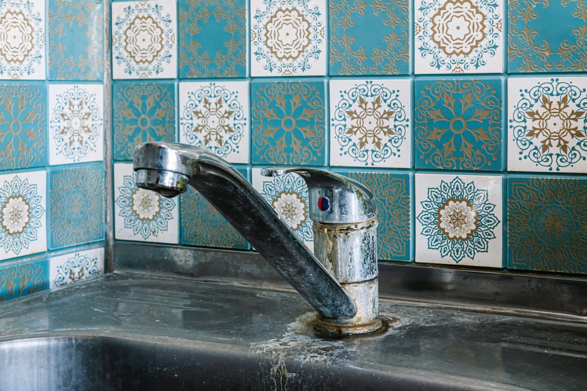 Close-up of an old-fashioned metal faucet over a stainless steel sink, in front of a decorative blue and white tile backsplash with intricate patterns, showing subtle signs of hard water stains.
