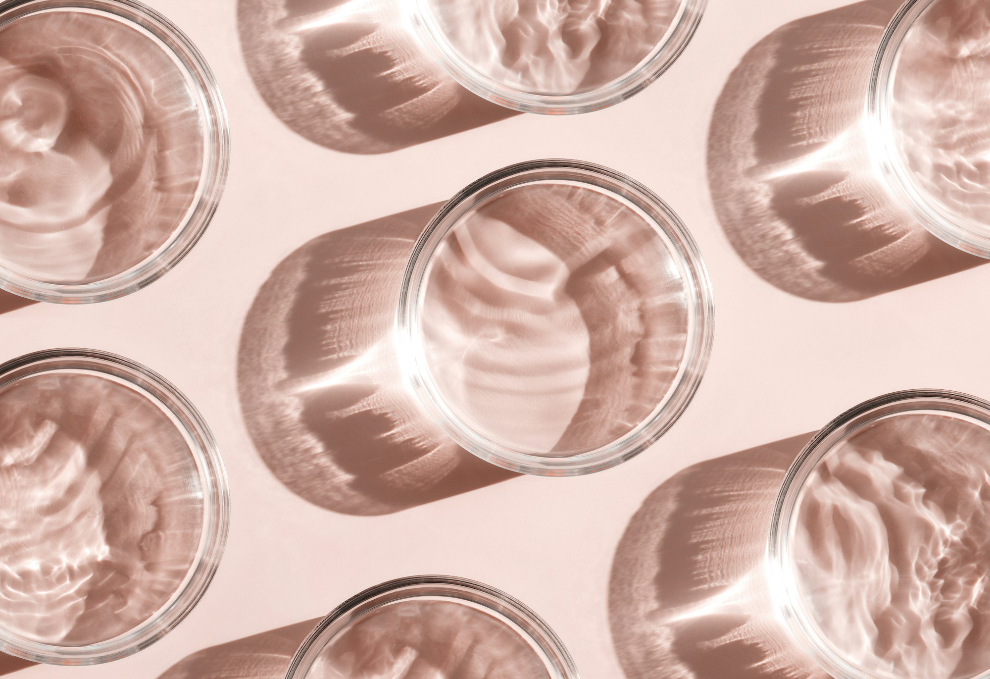Glass cups filled with water, casting complex, wavy shadows on a pink surface, arranged in a diagonal pattern—reminiscent of the ripples seen during diligent water softener maintenance.