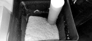 A container filled with granular material, possibly salt, features a white cylindrical object partially buried within the granules, hinting at the process involved in one of The 5 Stages of Water Softener Regeneration.