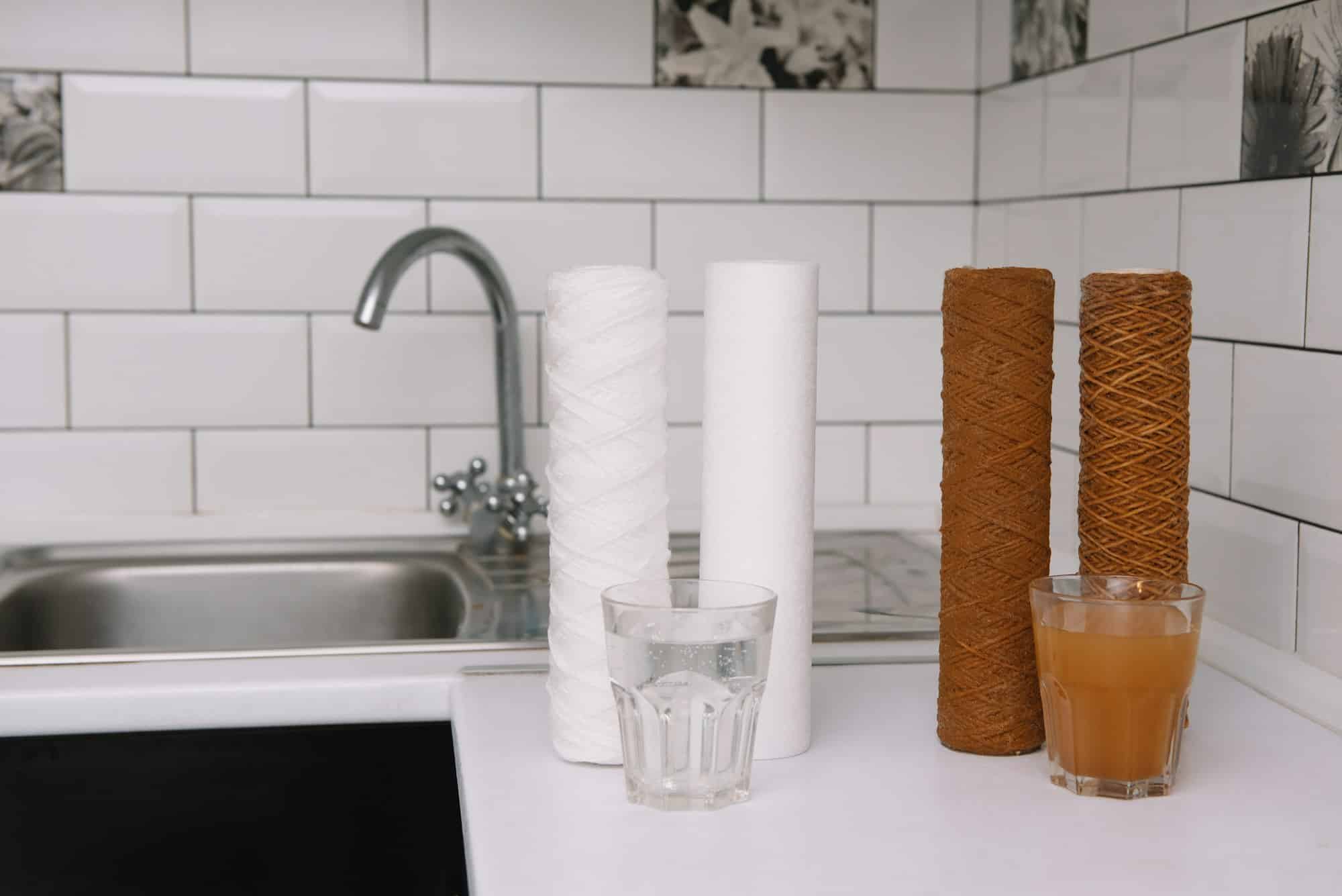 Two glasses of liquid are on a kitchen counter, one clear and one brown. Behind them are two white cylindrical objects and two brown cylindrical objects, likely for water softener maintenance. A tiled backsplash and sink with a faucet are in the background.