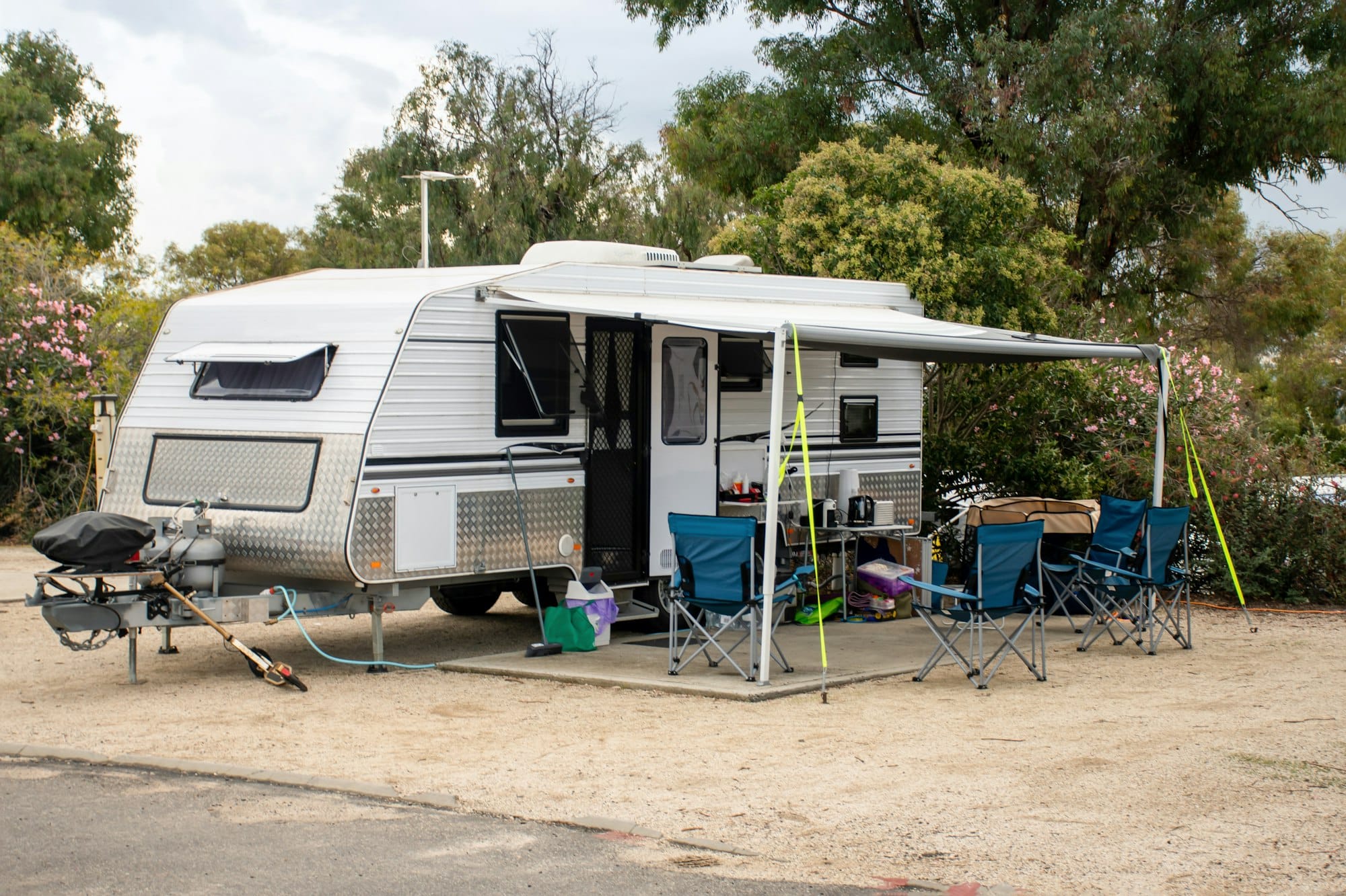 A silver travel trailer with an extended awning is set up at a campsite with several blue folding chairs and camping equipment arranged outside. Among the gear, the best RV water softener ensures fresh, clean water while trees and bushes surround the area.