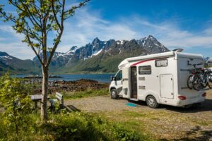 A motorhome is parked by a scenic lakeside with snow-capped mountains in the background. A tree and a picnic bench are in the foreground. Two bicycles are mounted on the back of the vehicle, perfect for when you take a break to install an RV water softener for fresh, clean water on your travels.