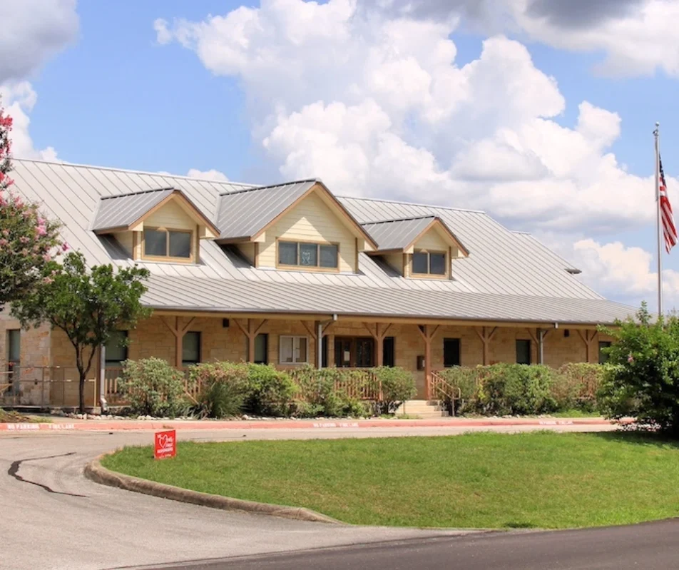 A large, two-story building with a metal roof and beige exterior stands as the Bulverde Texas City Hall. The well-kept lawn features a lamp post and two flags in the foreground, while trees and shrubs line the structure. A cloudy sky looms overhead.